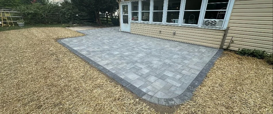 A recently installed stone paver patio in a backyard, featuring a smooth gray surface and bordered by landscaping. The image shows the patio area adjacent to a house with large windows, along with a straw-covered area and greenery in the background.