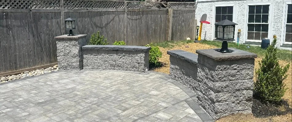 Patio area with stone pavers surrounded by short stone walls featuring built-in outdoor lanterns. In the background, there is a wooden fence, some plants, and a white house with large windows.