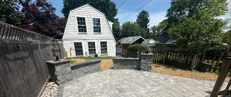 A residential backyard with stone paving, featuring a spacious patio area, bordered by a wooden fence and decorative stone walls, with a view of a white house and surrounding greenery.