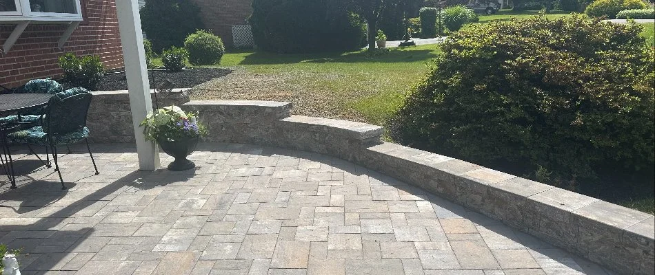 View of a stone patio dining area featuring a table and chairs, with a curved stone wall and landscaped surroundings, including shrubs and greenery in the background.