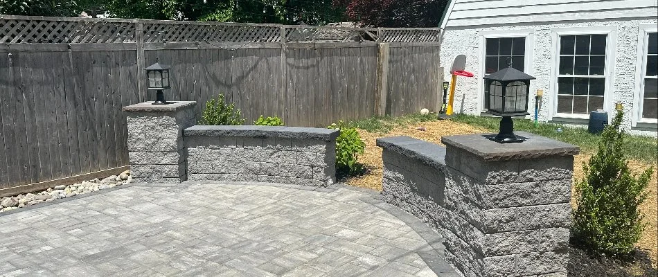 View of a stone patio area featuring curved stone walls with lanterns, surrounded by a wooden fence and a backyard with grass and landscaping elements